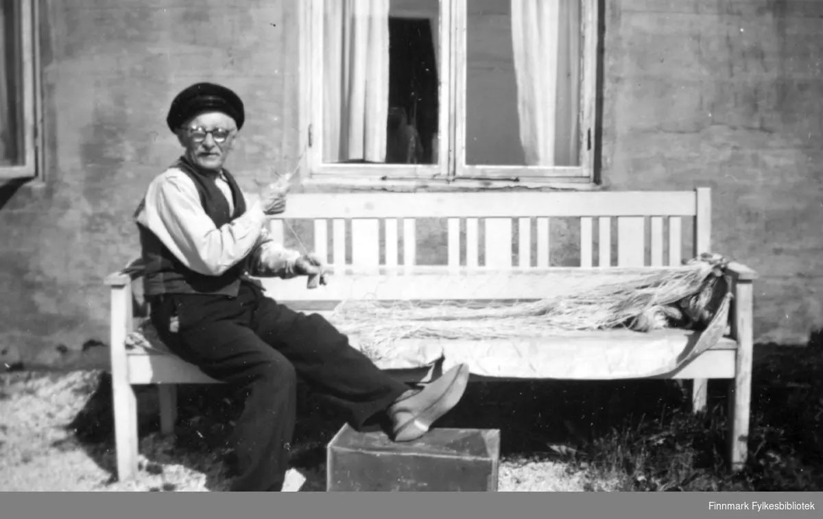Christian foran gamlehjemmet. Han sitter på en benk i sola og bøter garn Familiealbum tilhørende familien Klemetsen. Utlånt av Trygve Klemetsen. Periode: 1930-1960.