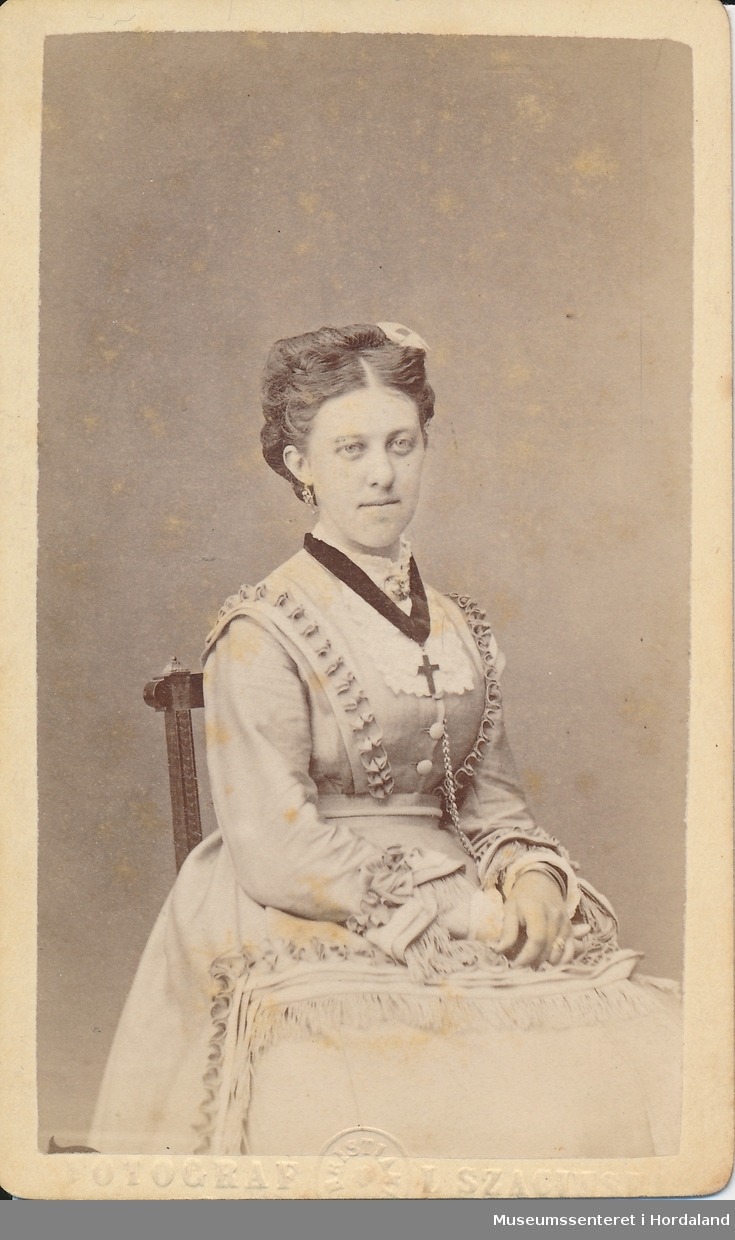 halvfigur fotografi av kvinne med midtskill, oppsatt hår, ljos bunadsliknande kjole med pynteband og halsband med kors 