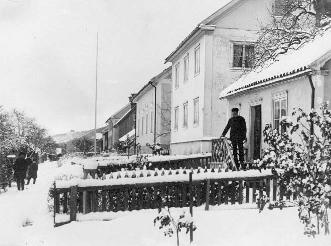 Bergsgatan i Gränna vintertid. Trädgårdsmästare Olsson står på farstutrappan till sitt hus. På gatan står okända personer vid en släde. I bakgrunden på höjden skymtar Brahehus.