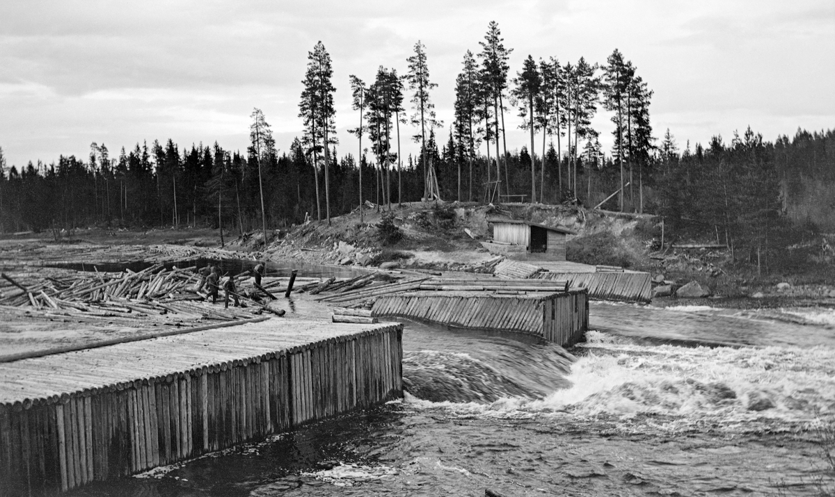 Tømmerfløting ved den gamle Kjellåsdammen i Flisa, ei sideelv til Glomma i Åsnes kommune i Solør. Denne dammen skal være bygd i perioden 1916-1922, med en kjerne av betong, forblendet med kavrete granittblokker. Da dette fotografiet ble tatt, i 1984, kan det se ut til at dammen var ytterligere forblendet ved hjelp av tømmer.  Kjellåsdammen hadde to om lag 11 meter brede løp, på hver sin side av et tømmerkledd kar, som var formet som et skråplan på motstrøms side.  Nettopp der hadde det samlet seg opp en del fløtingstømmer.  Fire karer arbeidet med å løsne de fastkilte stokker på dette stedet da fotografiet ble tatt.  På motsatt side av elva ser vi et borsdkur med pulttak nedunder en bakkekam der det vokste en del fine furutrær.

Kjellåsdammen var en såkalt atthaldsdam som kunne demme opp den nedre delen av Flisavassdraget i en lengde på opptil fem kilometer.  Den avbildete damkonstruksjonen ble erstattet av en som i stor utstrekning var bygd av kvadret naturstein i perioden 1916-24.  Det er nærliggende å anta at dette fotografiet må ha blitt tatt like før ombygginga.

Det første damanlegget ved Kjellåsfossen ble bygd på midten av 1800-tallet, jfr. fanen «Opplysninger».