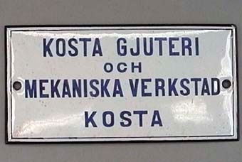 Rektangulär skylt av emaljerat järn med blå text på vit botten:
"Kosta gjuteri och mekaniska verstad Kosta".