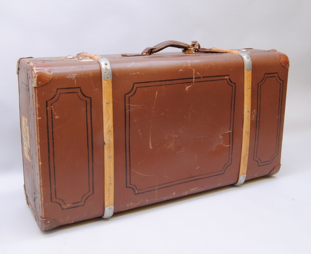 Brun resväska av papp med stomme av trä och två stycken ribbor av böjträ runtom. På väskans sidor är det målat ett dekorativt ramverk av svarta linjer likt speglar på dörrar eller väggpaneler. Resväskan har förstärkningar av metall i hörnen och låsanordning på var sida av handtaget.
Handtaget (:2) är av läder med en separat hållare för adresslapp med adressuppgifter tillhörande väskans ägare (se personuppgifter). På ena kortsidan sitter några lager av klistrade etiketter. På den översta etiketten går det att urskilja vissa ord: "Ankomst", "UMEÅ", "Till", "Över", "snälltåg", "Antal 2", "Tåg 2732". Den handskrivna texten är svår att läsa. På motstående sida sitter också flera lager av etiketter varav den översta är fullständig. Där står det "SJ Snälltåg", "Ankomstdatum:", "Från UMEÅ", Till: Eskilstuna", "Över: Vus.-Bä Sala-Kolbäck", "Antal kolli 3", "435", Totalvikt kg 50.", Tåg 2748", "Datum 4/1 1959". Längst upp i högra hörnet nedanför "Ankomstdatum" är det skrivet på snedden "129" samt "5/1".
Inuti är väskan klädd med mönstrat papper vars prickar och streck bildar ett rutmönster. I botten ligger ett flerfärgat mönstrat vaxat skyddspapper med rosa prickar och linjer. På insidan av locket sitter två gröna lappar som lagningar. Mellan lock och botten sitter bomullsband som håller locket i 90 graders vinkel från botten då väskan är öppen och bomullsband med spännen tvärsöver bottendelen för att hålla väskans innehåll på plats vid transport.