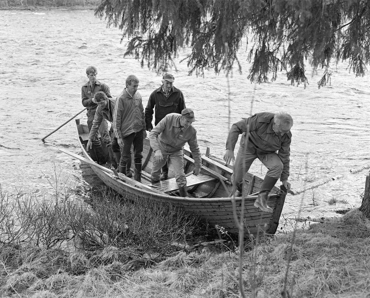 Båtutferd fra Stenbekkoia i Femundselva våren 1986, da styret i Klarälvens Flottningsförenings var på befaring langs den øvre delen av vassdraget. Bildet viser seks menn i en robåt som nettopp var lagt inntil elvebredden.  I akterenden sto det en «bakstavnkar» med ei skåtestang, som han brukte til å holde båten i posisjon mens de andre hoppet ut og inn på land.  Fløtingsbåtene som ble brukt i Trysilvassdraget i denne perioden var forholdsvis store, men likevel lette.  Båtbyggeren hadde brukt forholdsvis smale, tynne furubord, som han hadde sammenføyd ved hjelp av «kinger» (metallkramper).  De siste åra det var tømmerfløting i Trysilvassdraget var det arbeidsformannen Kåre Joar Graff som bygde fløterbåtene.  Dette handverket hadde han lært av onkelen sin, Ole K. Rømoe n, som også var en fløterveteran.  Da dette fotografiet ble tatt var det båtbyggern sjøl som satte seg til rette som «framstavnkar».