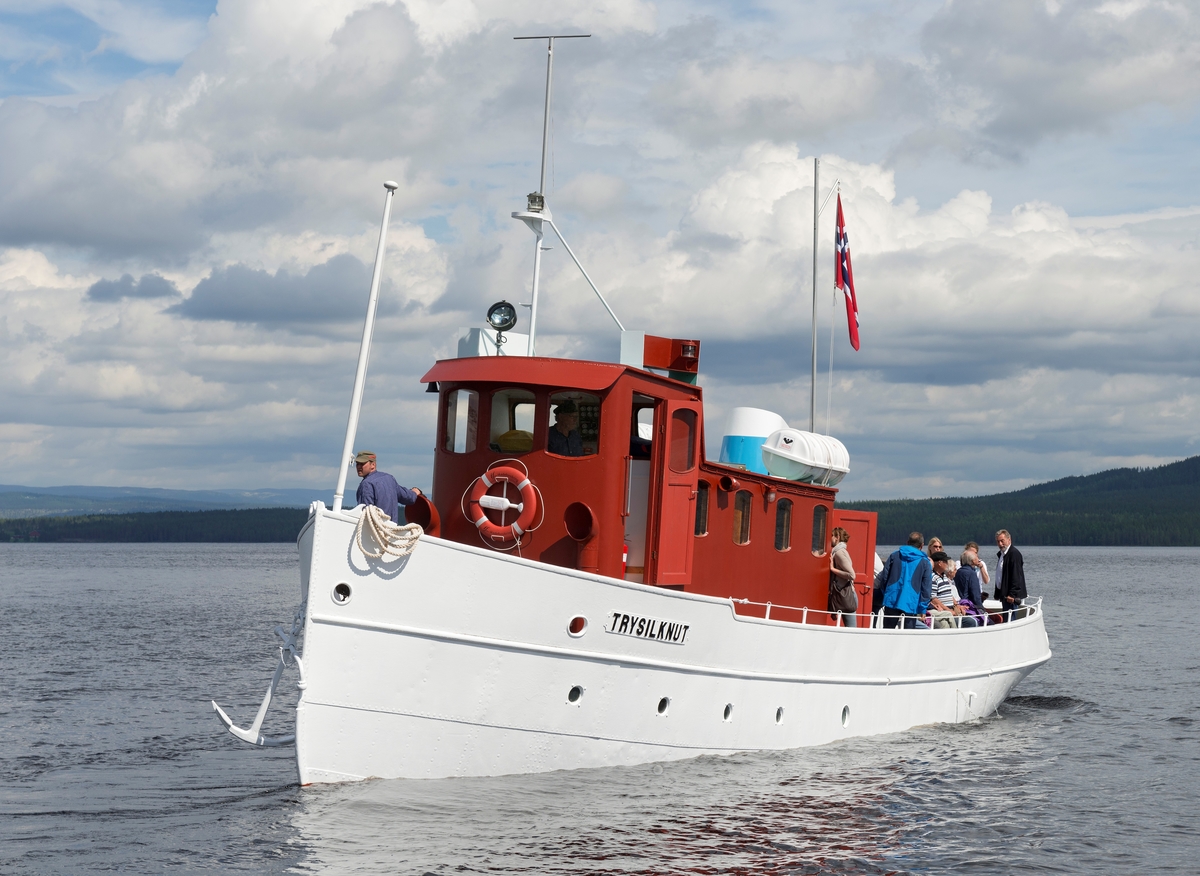 Slepebåten «Trysilknut» fotografert på den 47 kvadratkilometer store Osensjøen i grensetraktene mellom Åmot og Trysil i Hedmark, der Trysilknut gikk som tømmersleper fra 1914 til 1984.  Etter den tid har den 55 fot lange og 12 fot brede båten vært brukt som museumsbåt.  Den ble restaurert i perioden 2011-2014, og dette fotografiet er fra den første turen båten gjorde med passasjerer etter at restaureringa var avsluttet med maling av eksteriøret.  Skroget er kvitmalt og overbygningen (styrhus og bysse) er rødbrun.  Disse fargene har Trysilknut hatt siden båten ble ombygd fra damp- til dieseldrift i 1957-58, bortsett fra at vulsten som markerer hvor rekka møter den nedre delen av skroget lenge var grønnmalt.  Da dette fotografiet ble tatt sto skipper Størk Olsen til rors, og maskinist Allan Tutevdt befant seg på fordekket.  Passasjerene var gjester ved 100-årsmarkeringa for båten 11. juni 2014.  Blant dem skimter vi arkeologen Harald Jacobsen, direktør i Anno museum AS, lengst til høyre på akterdekket.  Mer om Trysilknut og dens historie under fanen «Andre opplysninger».