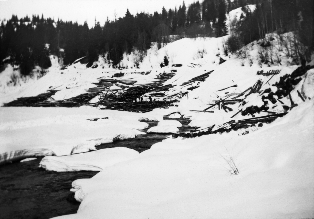 Fra Flatlandsvelta i elva Hjartdøla i Hjartdal kommune i Telemark.  Fotografiet er tatt vinterstid, på er tidspunkt da det var åpen råk bare over djupålen i vassdraget, mens landskapet for øvrig var dekt av snø.  I elveskråningen på nordsida av Hjartdøla aner vi en vegtrasé.  Fra denne vegen er det veltet sledekjørt tømmer nedover hellinga mot elveleiet.  Sentralt i bildet skimter vi en del karer som tilsynelatende har begynt å «florlegge» tømmeret i ei såkalt «strøvelte».  Dette innebar at stokkene ble lagt lagvis – i «floer» - med lengderetningen parallelt med strømretningen, og med mellomligggende «strøstokker» på tvers av strømretningen.  På denne måten oppnådde man å få ei luftig velte, der stokkene fikk en forsiktig tørk før våren kom og fløtinga tok til.  Dermed ble det noe mer lettflytende, og faren for at det sank og gikk tapt var betydelig redusert.  En annen fordel med slike strøvelter var at utislaget kunne utføres raskt og effektivt når vannføringa var ideell.  Tømmer som ble liggende hulter til bulter i elveskråningene derimot, var det besværlig å få slått uti, og fellesfløtingsforeningen kunne i enkelte tilfeller kreve refusjon fra skogeiere som hadde levert tømmeret sitt slik.