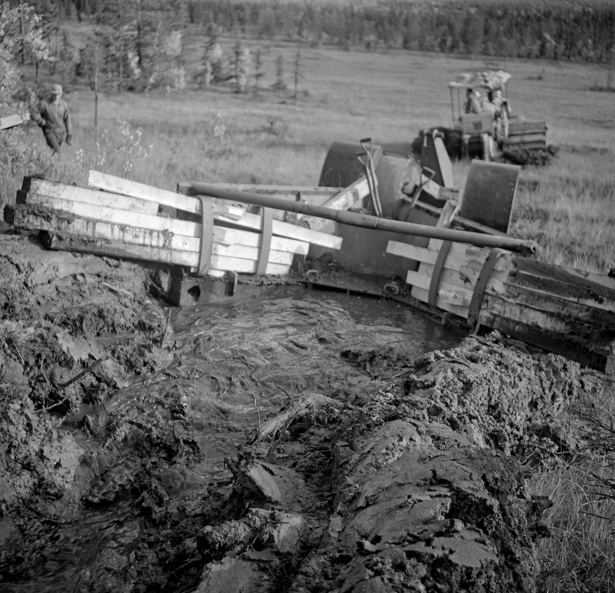 Såkalt «Lokomo-plog», fotografert bakfra under grøfteforsøk i Trysil høsten 1961.  Dette var en finskprodusert grøfteplog som veide hele 3,8 tonn.  Den ble trukket av en kraftig beltetraktor med bakmontert vinsj.  Lokomo-plogen etterlot seg ei renne i myra som var 70-90 centimeter djup, cirka 30 centimeter bred i botnen og en drøy meter bred i marksjiktet.  De oppløyde torvmassene skjøv plogen godt til side for grøftekantene.  Hvor langt vekk torva skulle skyves kunne justeres ved hjelp av plankestubber som ble tredd inn bak vertikale stålband på baksida av veltefjølene.  Da dette fotografiet ble tatt ble plogen kjørt på ei skrånende myr, med hellingsretningen.  Fotografiet viser også hvordan vannet fulgte plogen når den ble kjørt «unnabakke».  Mer informasjon om Lokomo-plogen finnes under fanen «Opplysninger».