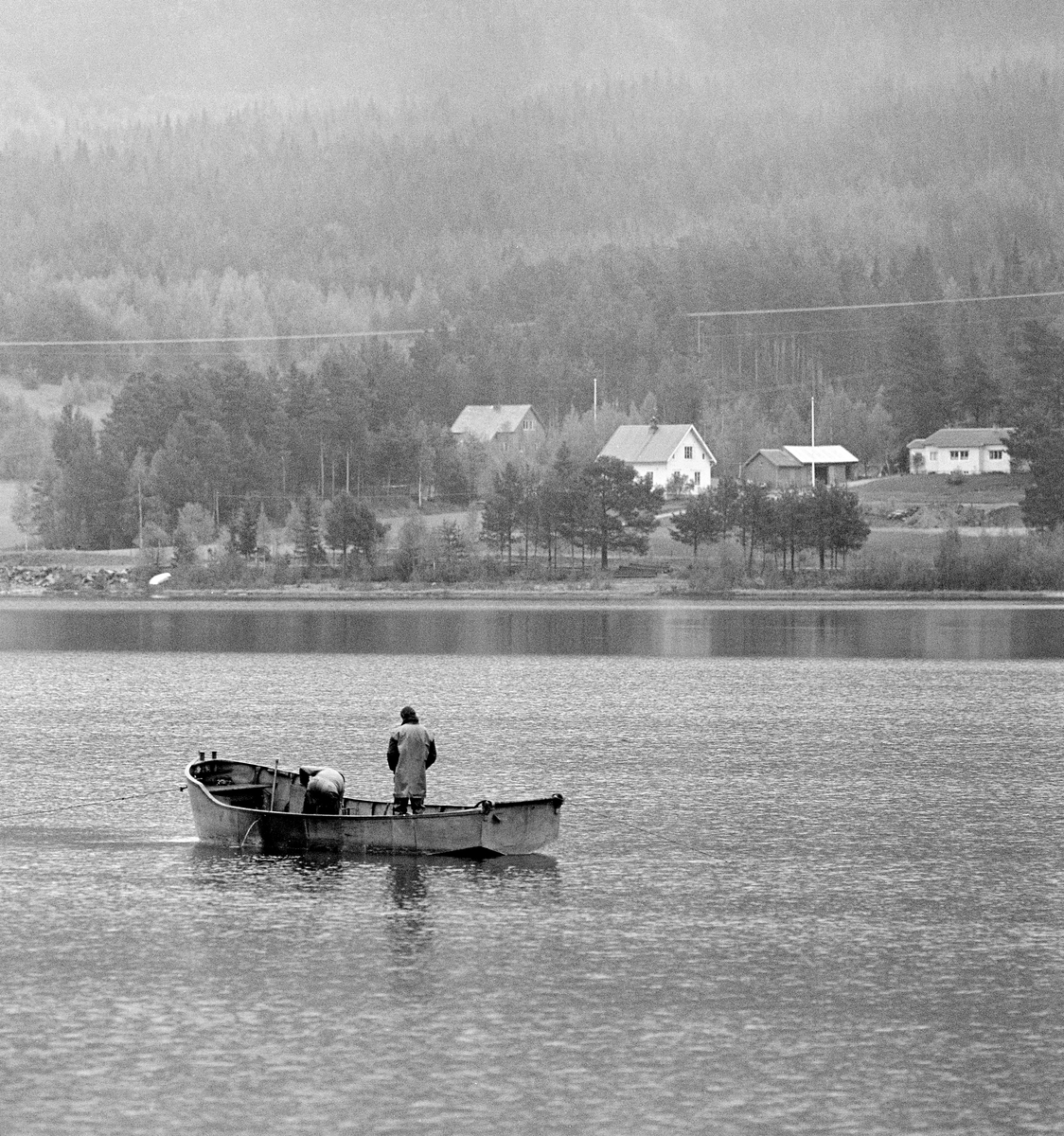 Tømmerfløting på Lomnessjøen i Ytre Renalen i 1981.  Fotografiet er tatt fra innsjøens vestre bredd, like nord for Kvernnesodden.  Ute på vannspeilet ser vi en av varpebåtene som ble brukt til å slepe ringbommer fra Hornsetlensa i nordenden hit til sørenden, der bommene ble åpnet slik at stokkene kunne flyte fritt videre nedover Åkrestrømmen mot neste innsjø, Storsjøen.  Båten hadde to manns besetning.