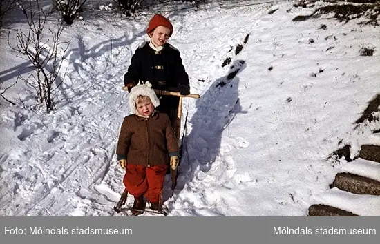 Två barn med en spark i snön. Det ena barnet kör och det andra åker med. 
Okända barn, plats och årtal.