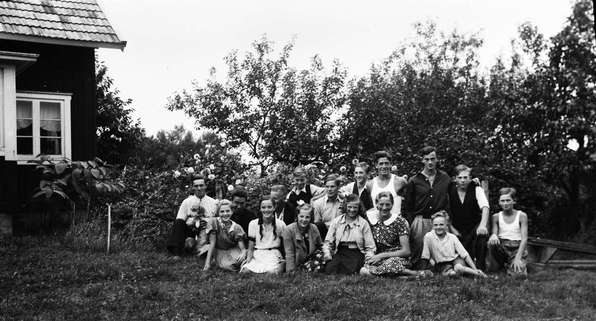 Sexton ungdomar från Drared sitter i trädgårdens gräs. Ynglingen längst till vänster håller en hund och pojken längst till höger är Gösta Andersson på Slätta. Det är sensommar eftersom dahlior blommar i rabatten bakom dem. Husknuten skymtar till vänster i bild.