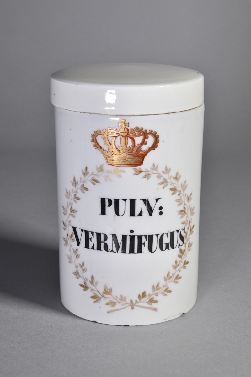 Ståndkärl, apoteksburk, av vitt porslin, cylindriskt med plant lock. Svart text inom guldfärgad lagerkrans krönt av krona.