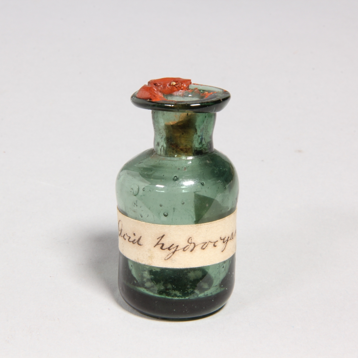 Flaskor, 10 st, av grönt glas med proppar av kork förseglade med rött lack. Står i pappask med lock klädd med marmorerat papper. Flaskorna innehåller rester av cyanid.