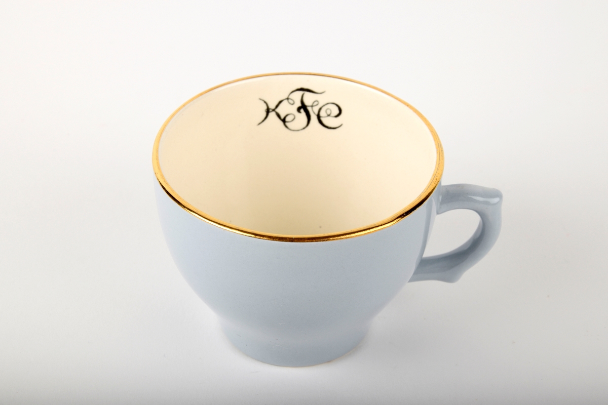 Kaffekopp med fat.

Koppen har monogram på innsiden. Både koppen og fatet har gullfarget kant. Fatet er hvitt i midten.