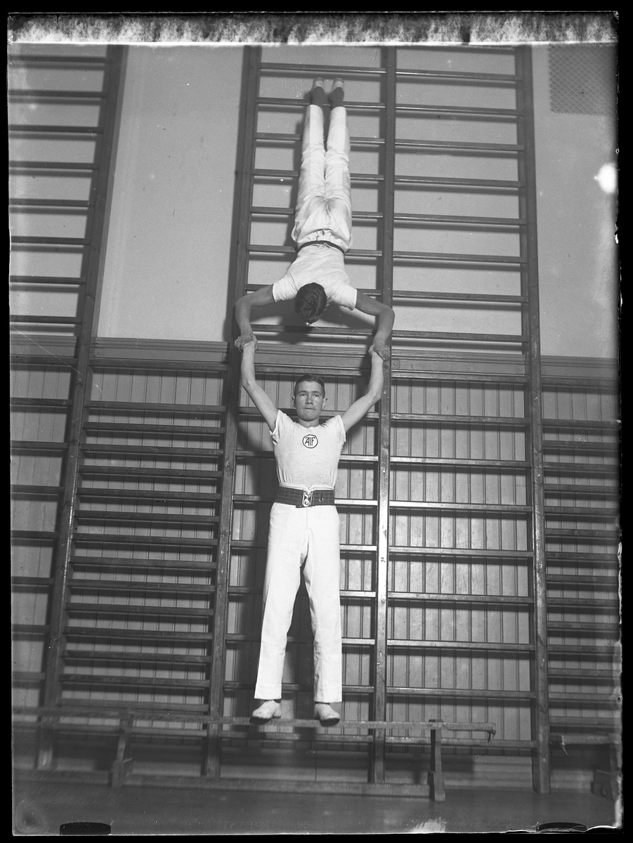 Hugo Larsson och Halvar Olsson i vita gymnastikkläder fotograferade vid ribbstol i gymnastiksal. Halvar lyfter Hugo som står på händer. Bilden är tagen för att användas i ett kollage (AM HO 00074).
