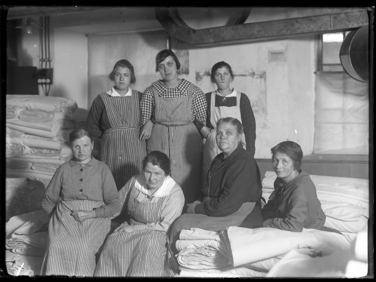 Sju av de arbetande kvinnorna i läggrummet på Alingsås Bomullsväveri. Fyra av dem sitter bland textilierna medan tre står arm i arm bakom.