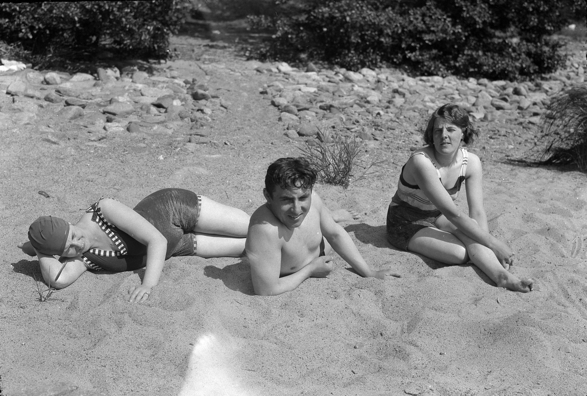 Två kvinnor och en man solar på en strand. Kvinnan som sitter upp är troligen Lizzie Skoglund. I Fotografens egna anteckningar står det "Besök på Risö, Badbilder".