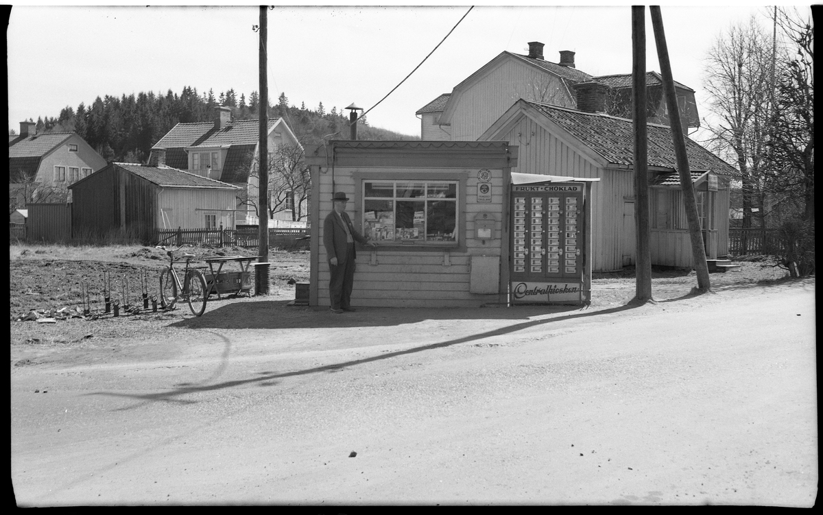 Centralkiosken på Boråsvägen 2, vid Malmgatan.
En liten kiosk av trä med liggande panel.
I bakgrunden flera boningshus i trä, olika tider och storlekar, även uthusbyggnader.

Framför kiosken alldeles till vänster om luckan står en man i kostym och hatt.

Till höger om kiosken står en väldigt stor varuautomat med många fack.
Över dessa står det FRUKT CHOKLAD och längst ner CENTRALKIOSKEN.

Till vänster står en cykel i ett cykelställ.

Stora telegrafstolpar.

I fonden Häradsberget.

Vägen mot höger är Boråsvägen i riktning mot Borås, Malmgatan går parallellt med järnvägen. Fotografen står ungefär där järnvägsövergången är.

1965 lades kiosken ned och huset flyttades till Saxebäcken för att bli sommarstuga.



text på negativkuvert:
Boråsvägen 2, april 1952
Boråsvägen 25, maj 1957
Innehavare Gunnar Risberg.
Kiosken flyttades 28.6.1965 till Saxebäcken, för att användas som sommarstuga.
Här ligger kiosken i hörnet av Boråsvägen och Malmgatan. När är då bilden tagen? Jämför med andra, daterade, bilder av kiosken.