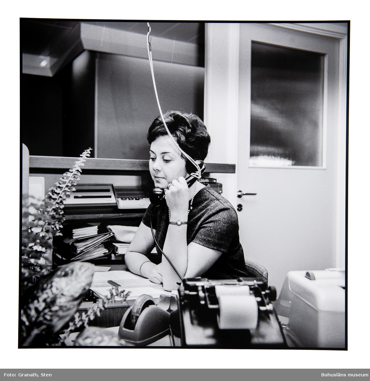 Växeltelefonisten Anita Fransson på Domus sitter vid ett skrivbord och talar i en telefon som hänger i ett snöre från taket.