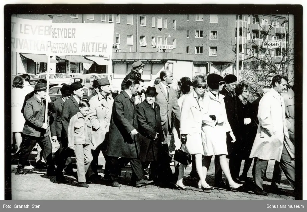 Demonstrationståg längs Göteborgsvägen vid Folkets Hus. Några personer håller i en banderoll med texten "[...]t lever[...]betyder [...]rivatkap[...]alisternas aktier". I tåget, som rör sig mot höger i bilden, går kvinnor och män i alla åldrar. I bakgrunden Almqvist´s Korvkiosk och Asplundsgatan.