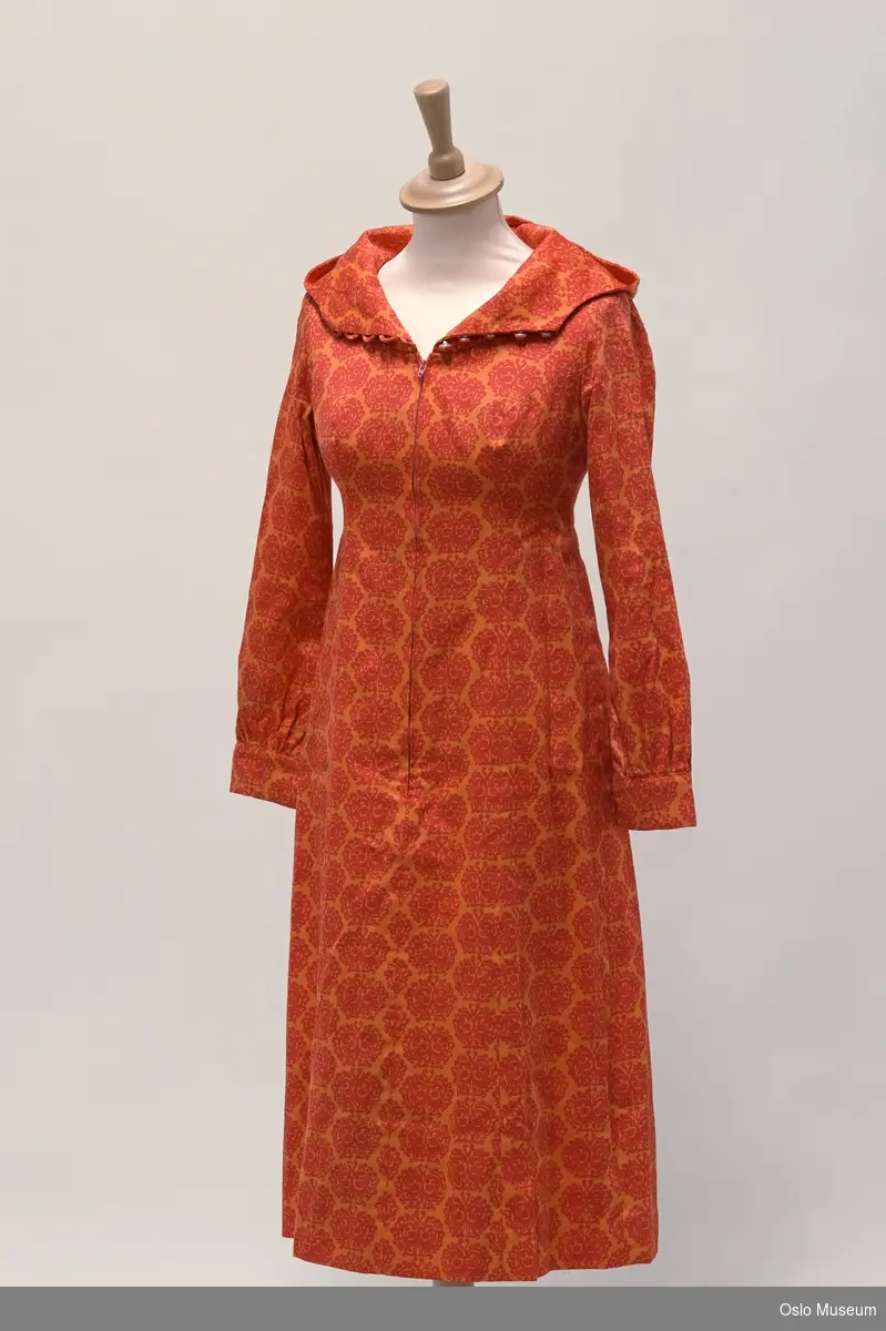 Sid kjole med hette og knapper og glideklås foran.
Bomull i oransje og rødt blomstret mønster.