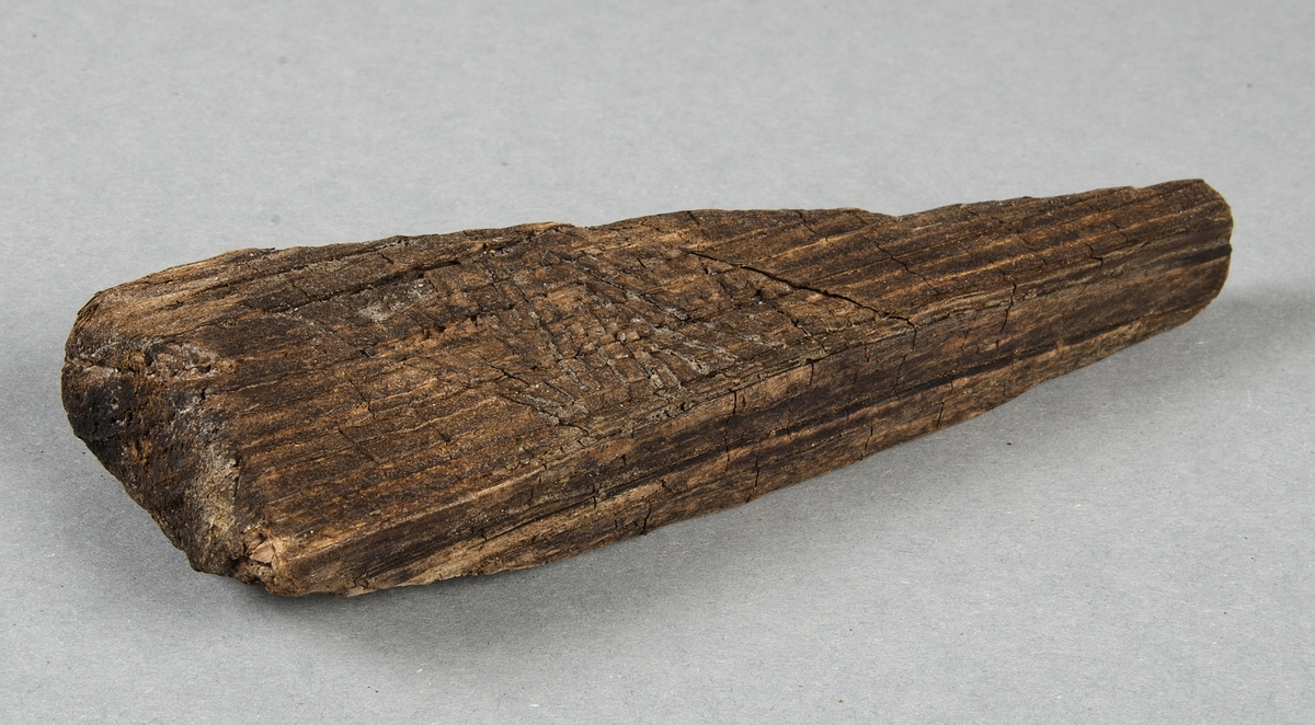 Bearbetat trä. Triangulärt föremål med rektangulärt tvärsnitt, trolig möbeldetalj. Dekorerad med linjeornamentik.