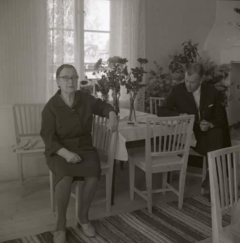 En man och en kvinna sitter på varsin stol intill ett bord. I bakgrunden syns en öppen spis, 1968.