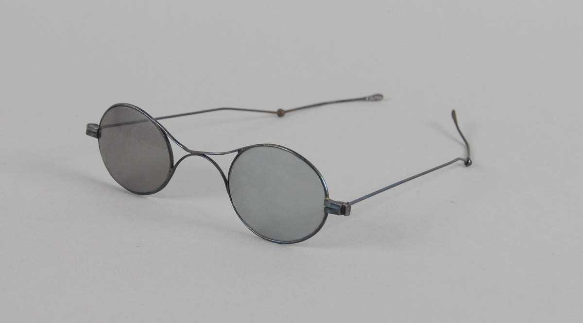 Ovale briller med mørkt glass og metallinnfatning. Stengerne kan brettes ut.