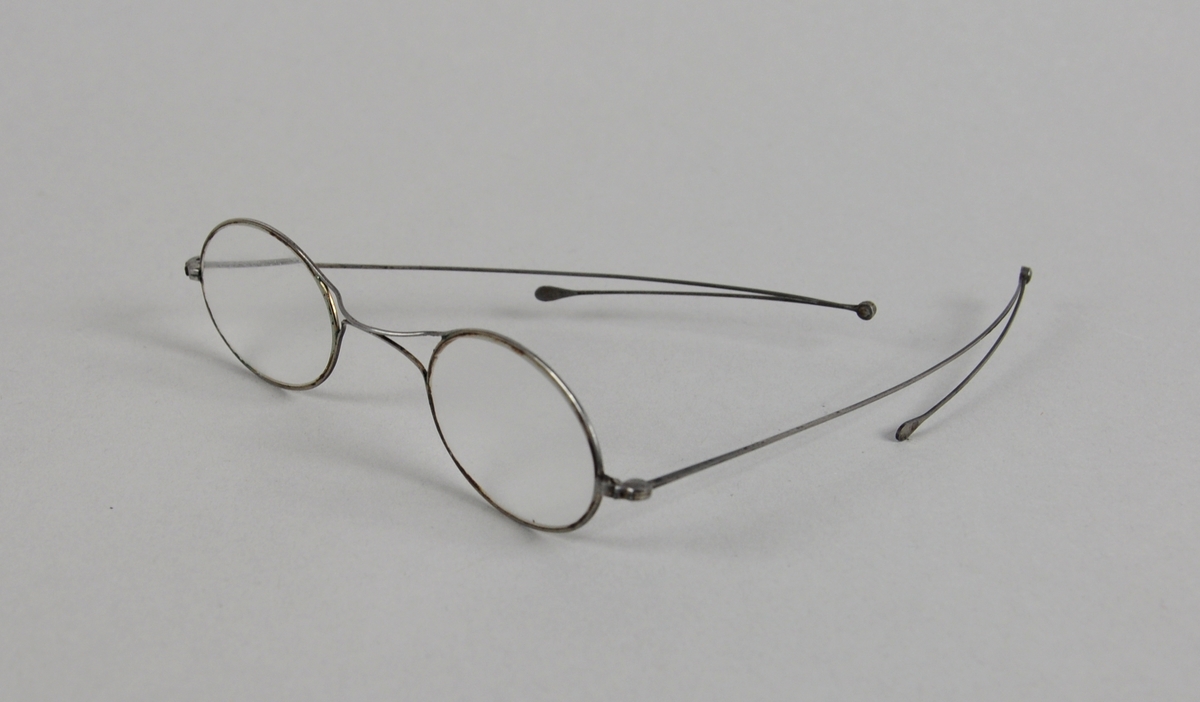 Ovale brilleglass med tynn metallinnfatning. Ytterste ledd på brillestengene kan brettes ut.