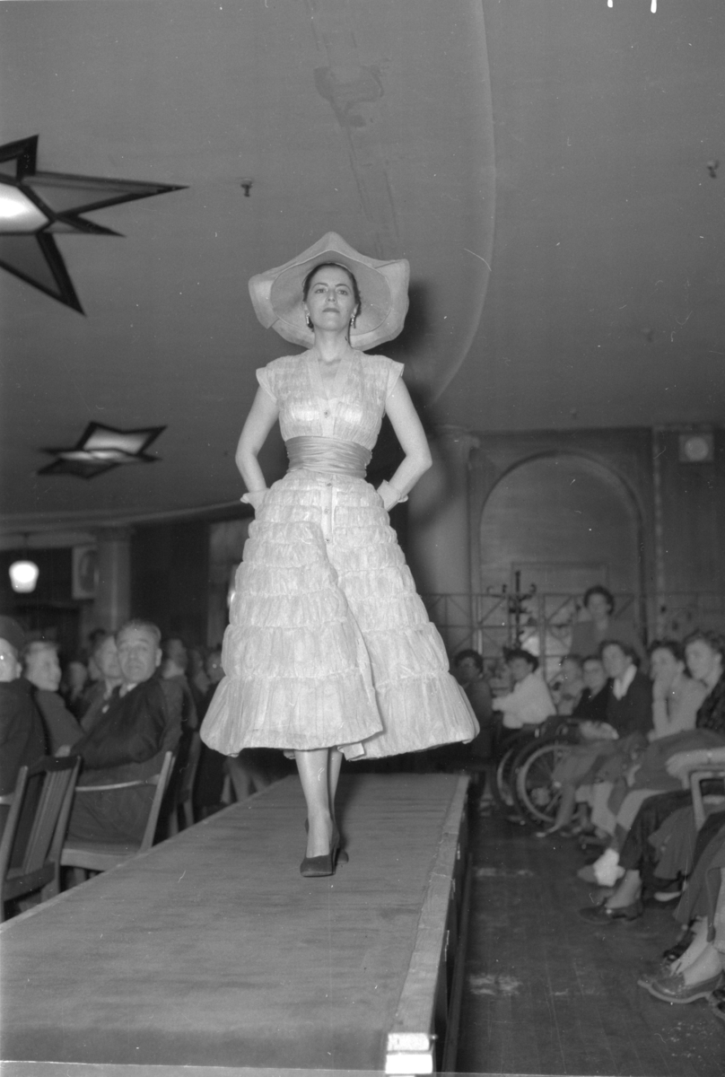 En kvinne på podiet iført kortermet kjole og en stor hatt. Fra en motevisning på Steen & Strøm fra 7. mai 1954..