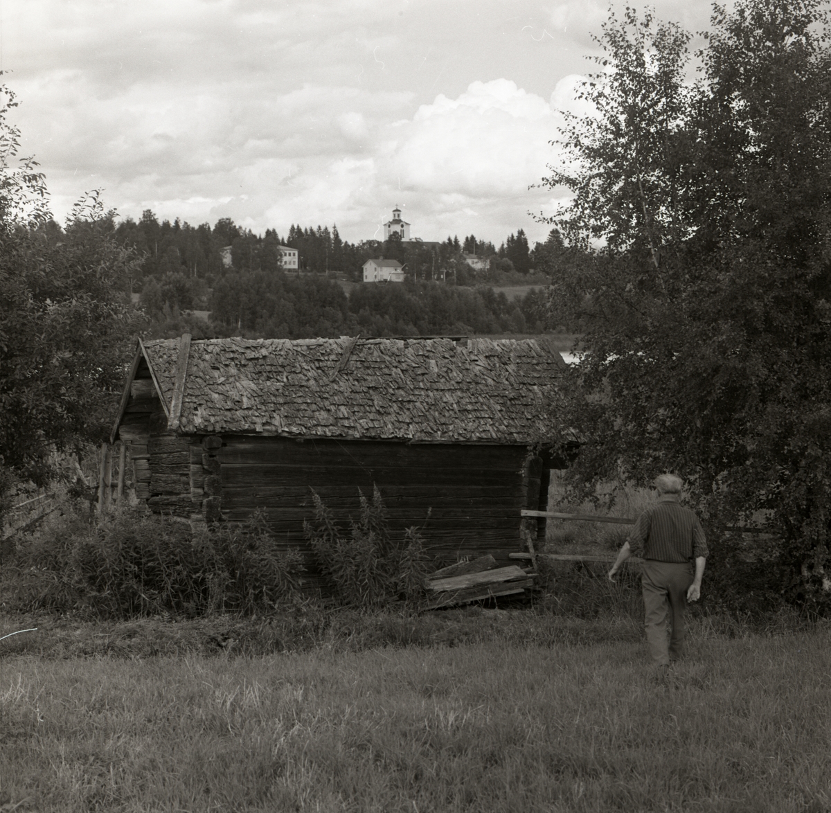 En man promenerar mot en lada med spåntak, i bakgrunden syns Rengsjö kyrka samt bostadshus, 1972.