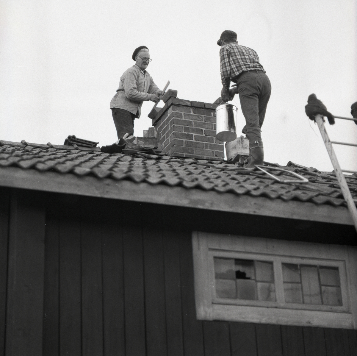 Två personer står på ett hustak och murar en skorsten i Rengsjö, 23 maj 1975.