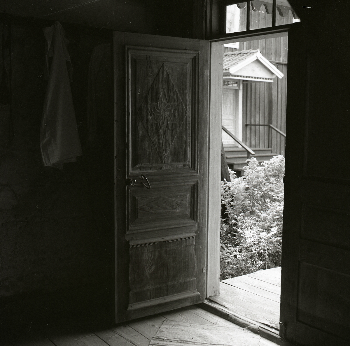 En trädörr med snidade dekorationer står öppen och utanför syns en förstukvist med snickarglädje, handtag, tröskel och överljusfönster. Innanför trädörren finns ett trägolv med en trasmatta och en jacka hänger bakom dörren, Hembygdsbyn 1 september 1981.