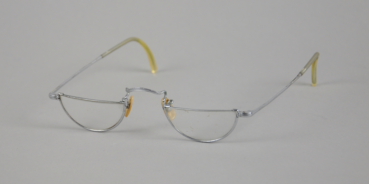 Briller med halvmåneformede brilleglass og metallinnfatning. Plastbelegg ytterst på stengene.