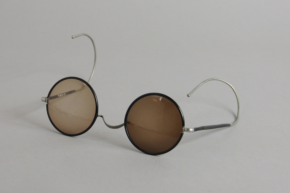 Runde mørke solbriller med metallinnfatning. Rundt brilleglassene og den innerste delen av stengene er forsterket med svart plast. Det høyre brilleglasset er mørkere enn det venstre.