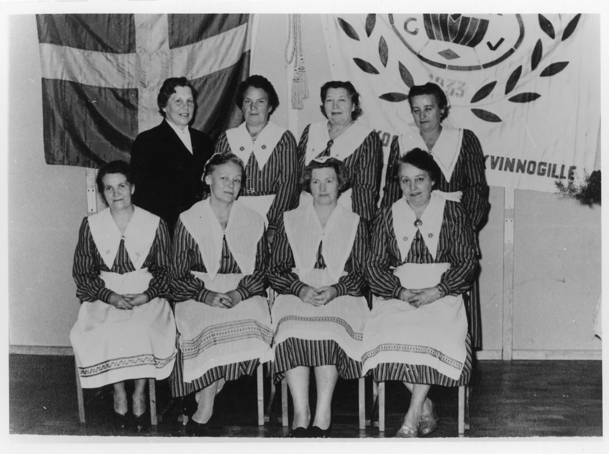 Gruppbild av 8 kvinnor från styrelsen av Kooperativa Kvinnogillet varav fyra står och fyra är sittande. I bakgrunden ses svenska flaggan och deras organisationsflagga. Bilden är tagen under deras 25-års jubileum, 1933-1958.