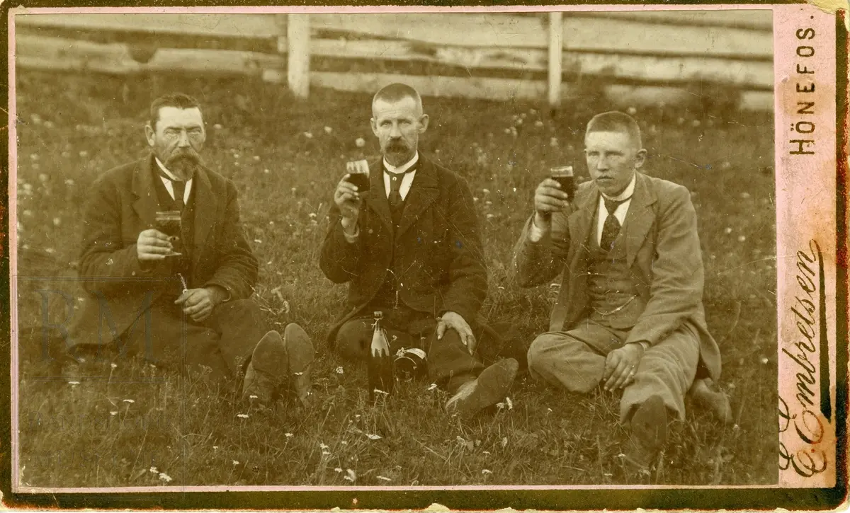 Tre menn sitter i gresset og skåler. Fra venstre Kristian Andfossen, Julius Henriksen og Hans Kristian Andfossen.