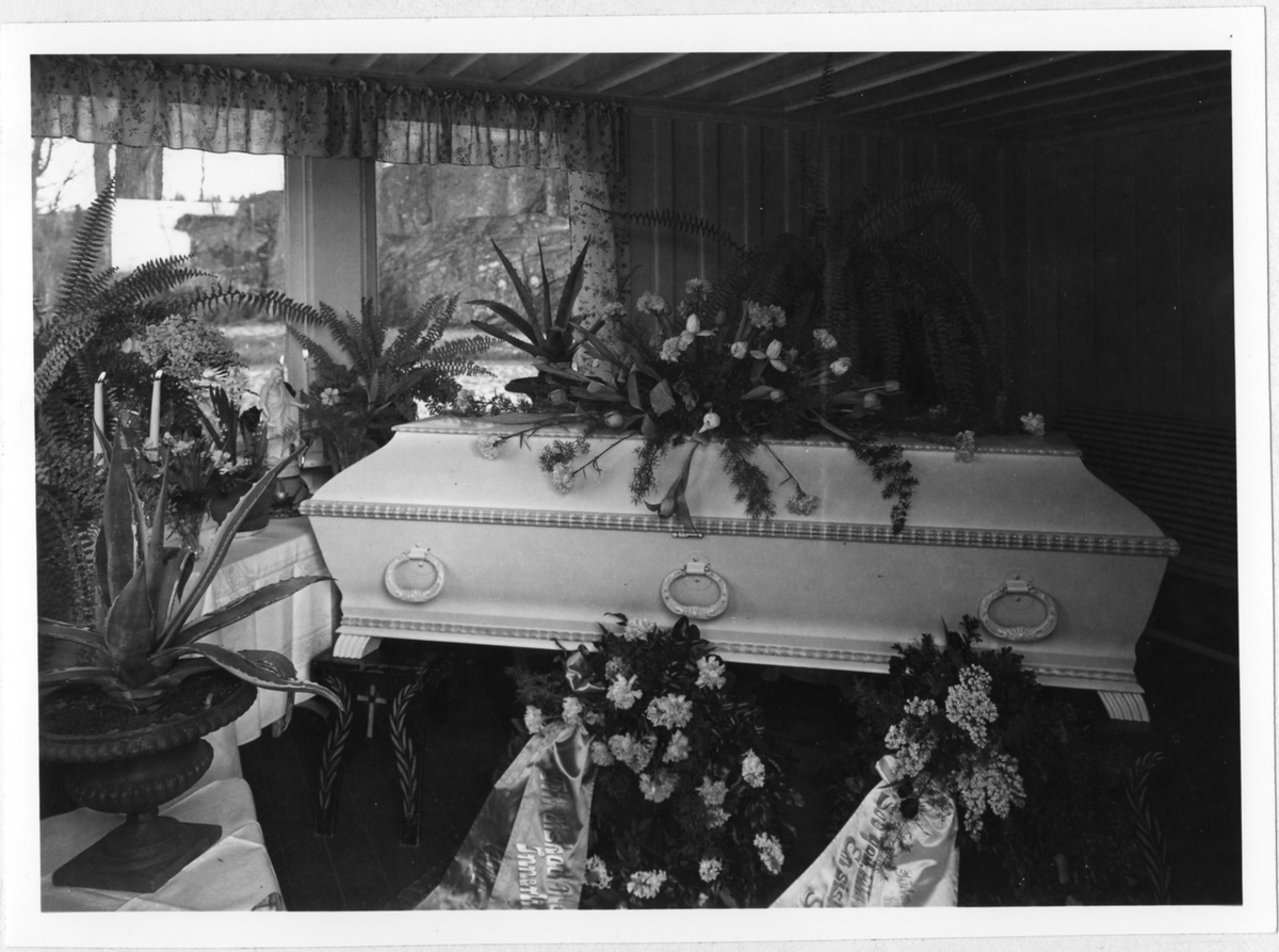 En kista täckt med begravningsblommor, en liten statyett av Jesus står i fönsterkarmen.

Begravning år 1947 i Alingsås.