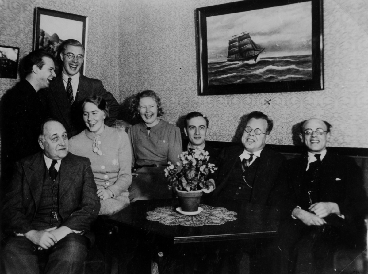 Gruppbild av 6 män och 2 kvinnor som skrattande trängs på en soffa. På väggen bakom dom hänger tavlor, där ena har motiv av ett skepp till havs. 
Esperantoklubben i Alingsås. Mannen längst till höger är Erik Edholm.