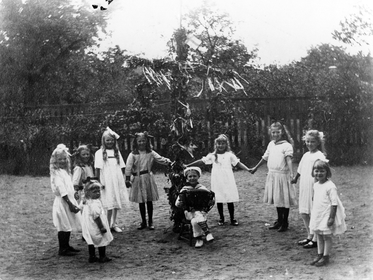 Nio stycken flickor dansar kring en midsommarstång och en liten pojke sitter vid stången och spelar dragspel.