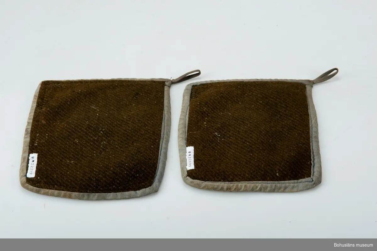 Ett par slitna grytlappar av kypertvävt ylletyg kantade med bomullsband.
Möjligen tillverkade av äldre ytterplagg.