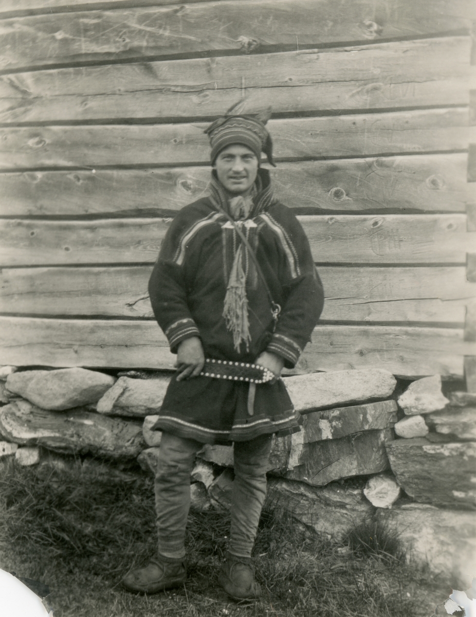 Portrettfotografi av en samisk mann ved navn Hætta i samekofte ved ei stove i Bingis, Kautokeino.