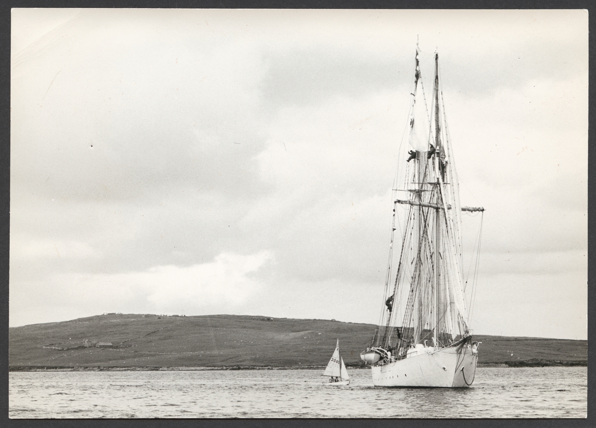 Marinens skolfartyg Gladan ligger till ankars utanför Falmouth England. Fartygsporträtt framifrån med hissade segel. Från styrbordssidan närma sig en slup som seglas av två personer. I bakgrunden syns Cornwalls kust.