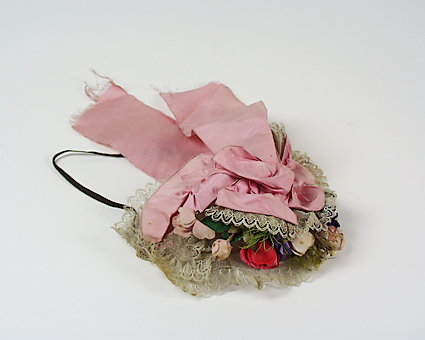 Hårklädsel, ungmorsmössa, av vit tyll med spetskanter och konstgjorda rosa, röda och blå blommor och gröna blad och fjädrar. Rosa sidenband i nacken. Nacksnodd av gummiband. Ståltrådsställning.