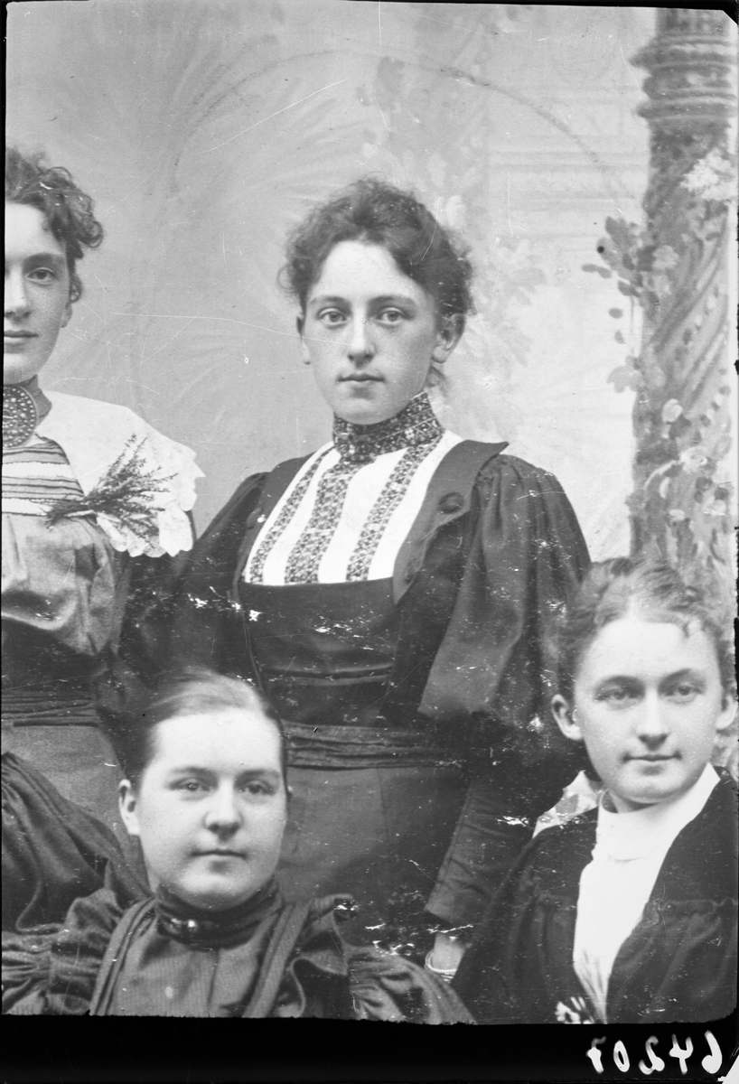 Gruppebilde. Gruppe på fire kvinner. Kopi av positivt foto. I senter sitter en ung mørkhåret kvinne med oppsatt hår i en mørk kjole med lys front med pynt. Bestilt av Ludvig Eriksen