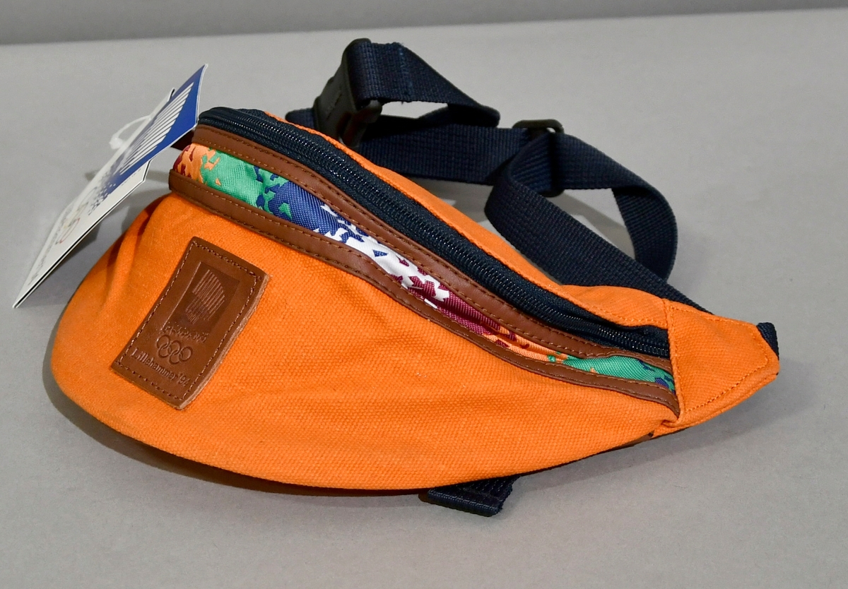 Oransje rompetaske med glidelås og skinnkanter. Under glidelåsen en stripe med krystallmønster i grønt, blått, rosa, hvitt og oransje. Skinnmerke med emblemet for Lillehammer '94. Ekstra lomme med glidelås på innsiden.