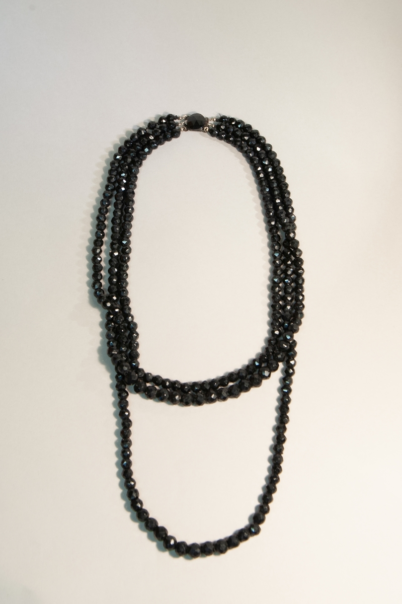 Halsband av svarta stenkolspärlor. Slipade kulor uppträdda på silkestråd i tre rader av varierande längd. Lås i vitmetall med en slipad stenkolsplatta.