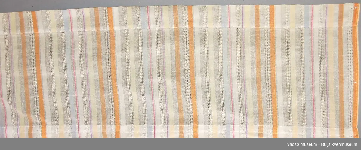 Stripemønstret gardinkappe i tynt linstoff. Vertikale 0,5-2 cm brede vekselsvise striper med transparent, heklet hvitt, blått, rødt og oransje.