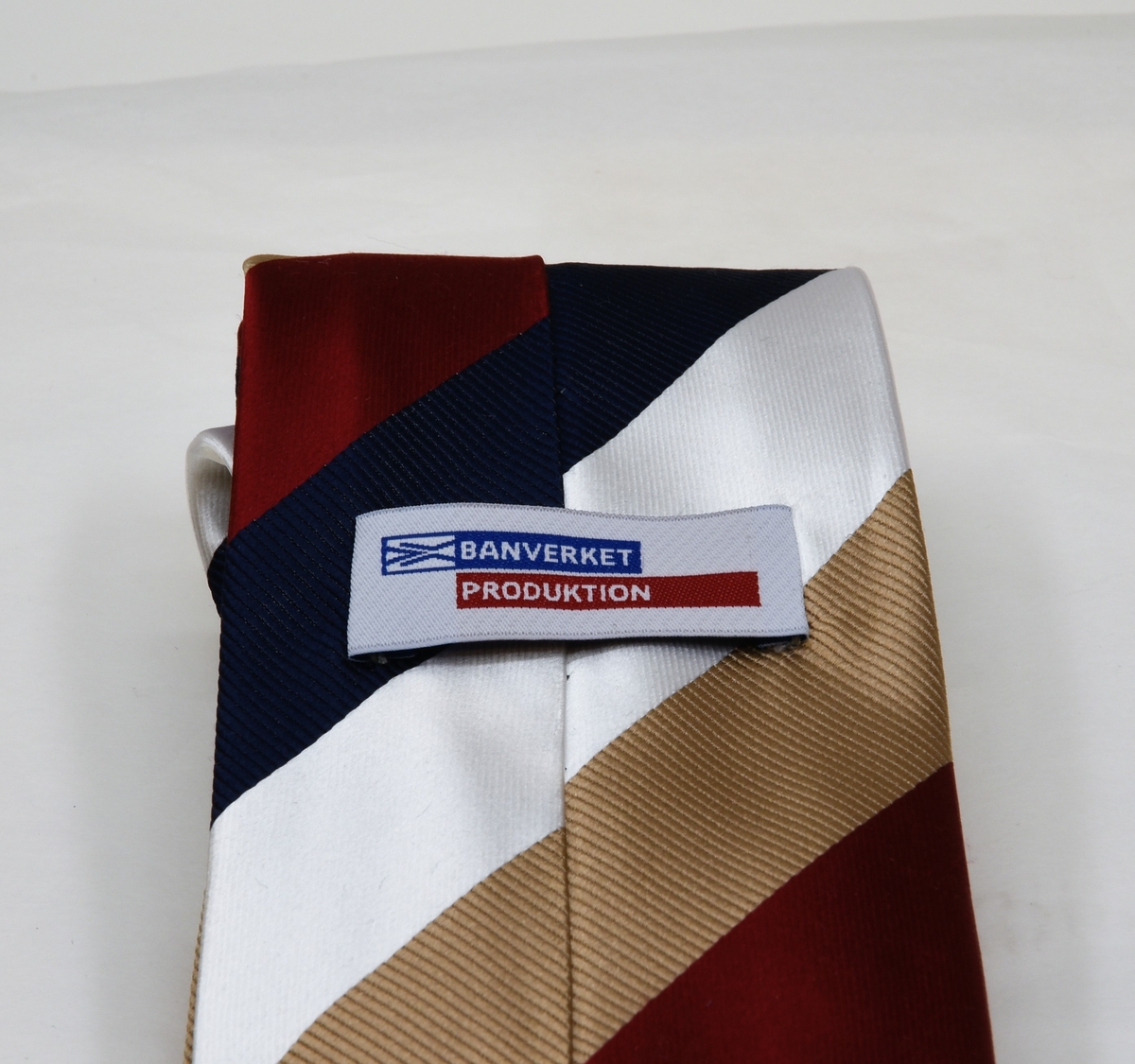 Slips av silke med diagonalrandigt mönster i färgerna vit, blå, röd och ljusbrunt. På baksidan av slipsen sitter det två lappar.