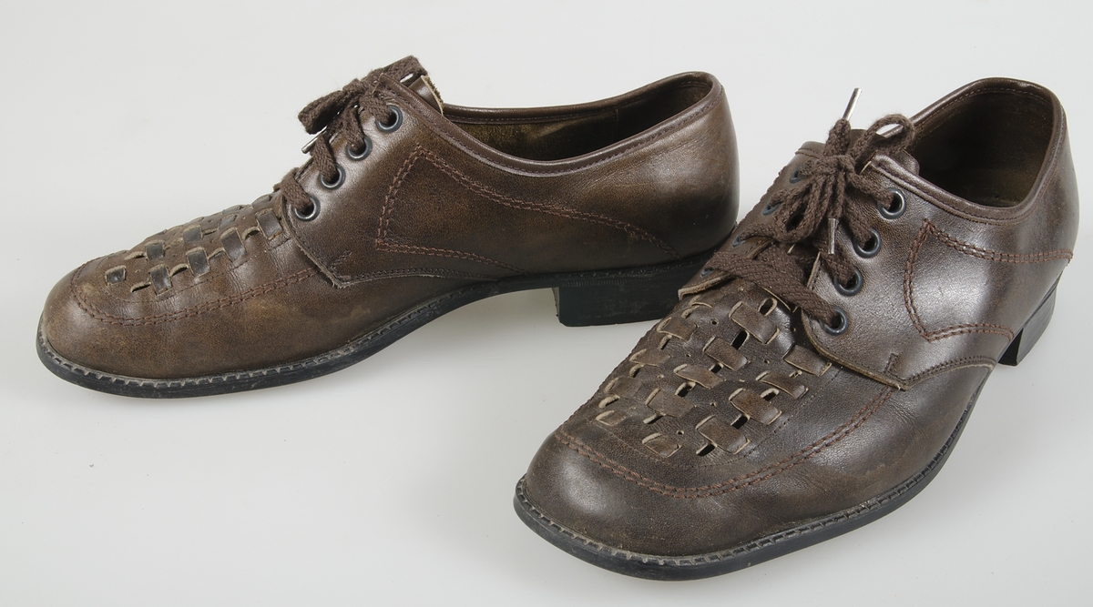 Ett par snörskor av mörkbrunt läder med flätad ovansida. Mörkbruna snören. Åtta snörhål på varje sko. Gummisulor utanpå. Träsulor inuti. Maskinsydda. Räfflade klackar. Storlek 7½. Märkt: AA. Samhörande med nr. UM 19115-19188.
