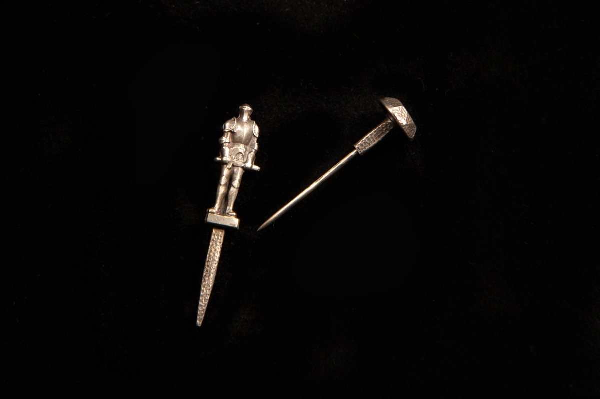 Kråsnål av metall i form av en stående riddare i rustning med svärdsbalja bakom. På nålens huvud ingraverat en riddare i rustning med årtal på var sida: "19 15".