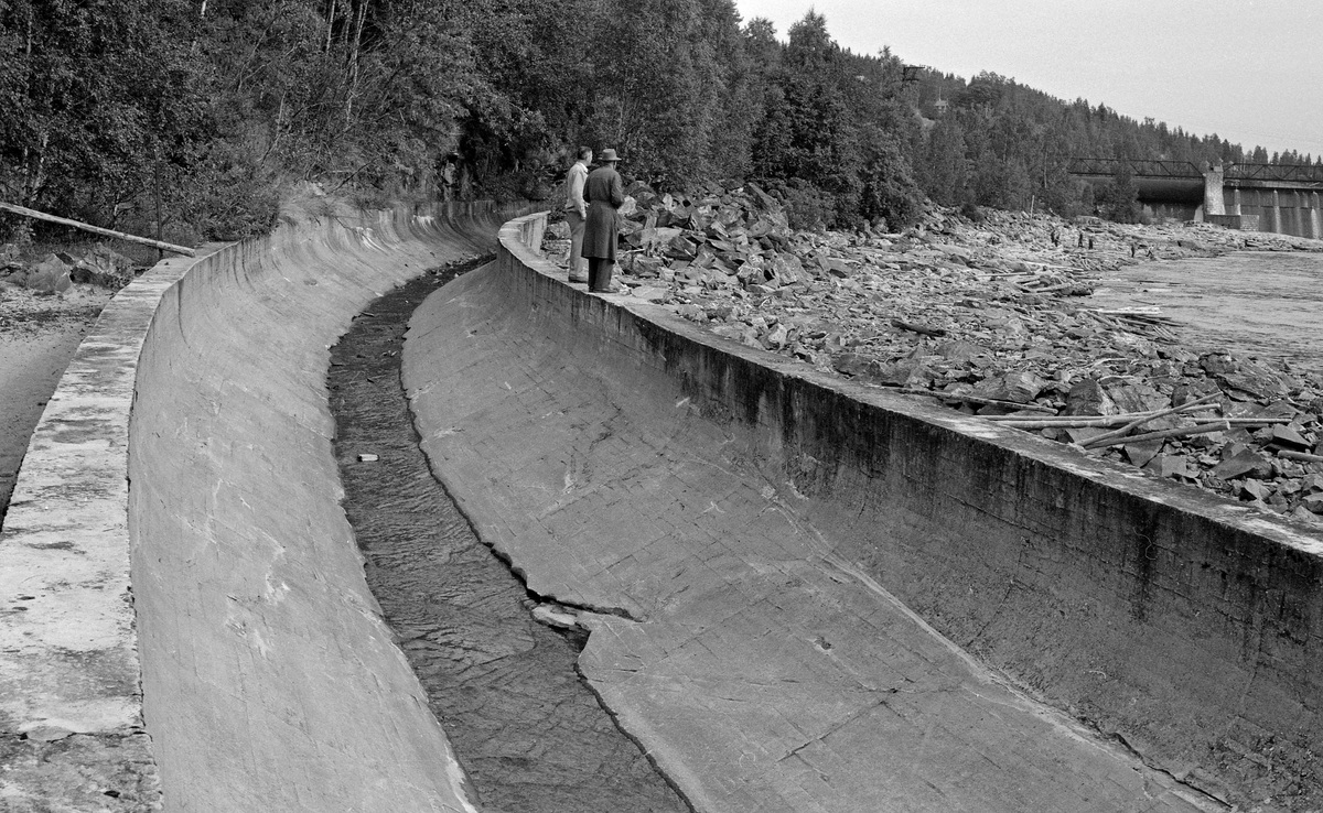 Tømmerrenna ved Rånåsfoss kraftverk i Glomma.  Dette anlegget, som ligger i Nes kommune i Akershus, ble bygd for Akershus fylke i perioden 1918-1922.  Inntaksmagasinet til dammen har et areal på cirka 10 kvadratkilometer.  Det strekker seg over to mil oppover i elveløpet, nesten til Svanfossdammen og Funnefoss.  For å økonomisere best mulig med vannreserven også i tømmerfløtingssesongen ble det bygd ei renne langs den vestre elvebredden.  Denne fikk en lengde på cirka 800 meter.  Den øvre delen gikk i en fjelltunnel, som ble foret med betong, slik at stokkene ikke skulle kile seg fast.  Den nedre delen av renna derimot, hvor dette fotografiet er tatt, er en åpen betongkonstruksjon, støpt med et U-formet tverrsnitt.  Rånåsfossrenna ble konstruert med henblikk på at den skulle kunne ta unna om lag 180 000 tømmerstokker per døgn med ei vannføring på 15 til 30 kubikkmeter i sekundet.  Framfor renneinntaket i kraftverksdammen ble det bygd en 15 meter bred «inntaksskuff». Det viste seg imidlertid å være ei bakevje ved renneinntaket, noe som gjorde påstikkinga svært mannskapskrevende. Følgelig ble mesteparten av tømmeret i stedet sluppet gjennom ei av de to sektorlukene i Rånåsfossdammen. Under høykonjunkturen som preget norsk skogbruk i begynnelsen av 1950-åra kunne det gå opptil 700 000 tømmerstokker forbi Rånåsfoss i høysesongen for tømmerfløtinga. Da dette fotografiet ble tatt – sommeren 1961 – sto to fløtingsfunksjonærer på rennekanten og betraktet det tømmeret som hadde strandet på den steinete elvebredden. Oppi renna lå det en enslig stokk som det sildret litt vann under.